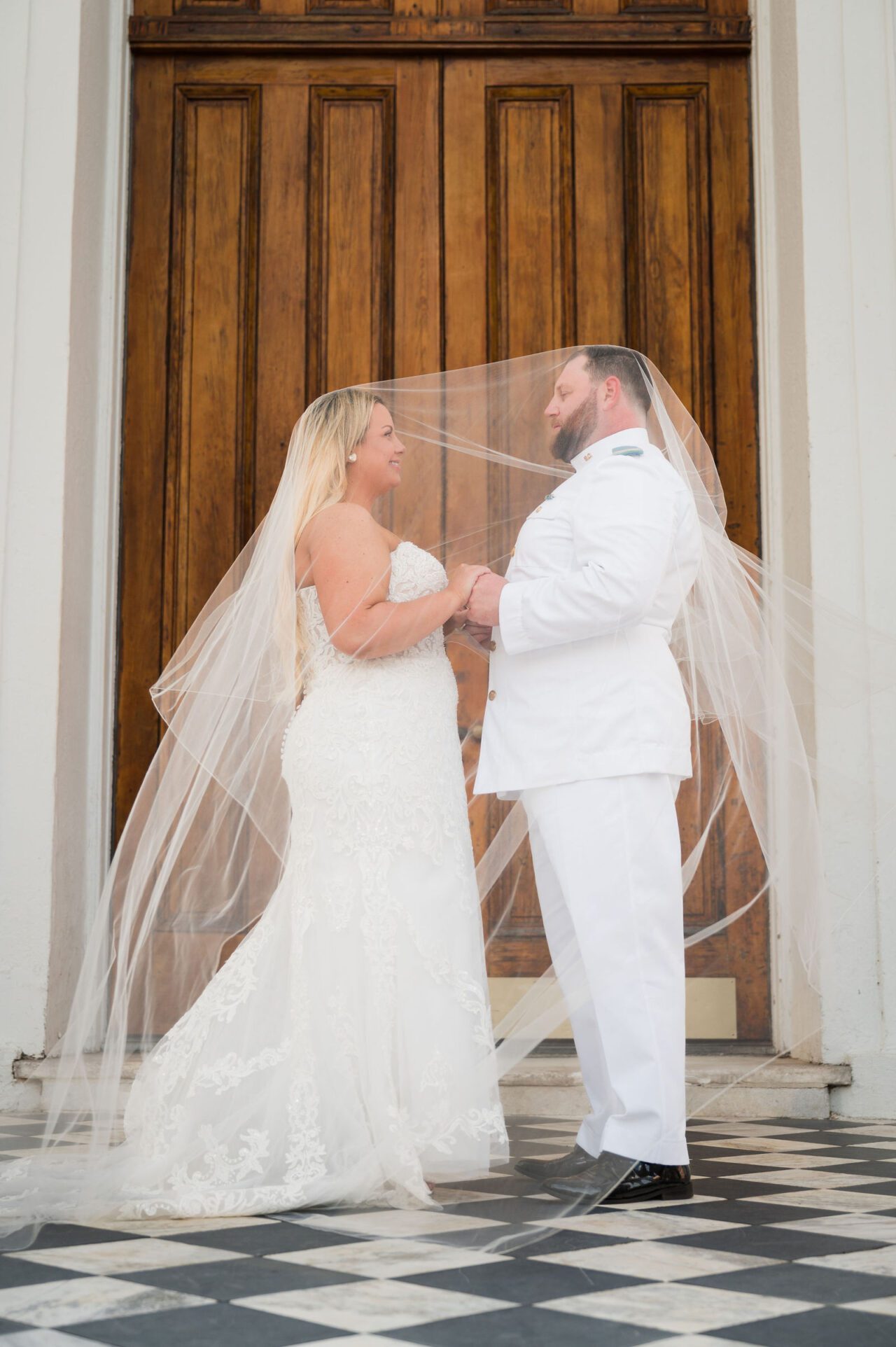 Bride and groom sharing a romantic wedding portrait beneath a flowing veil in downtown Charleston, South Carolina, with elegant wooden doors and classic black and white flooring. Timeless Charleston wedding photography with cinematic and editorial style.