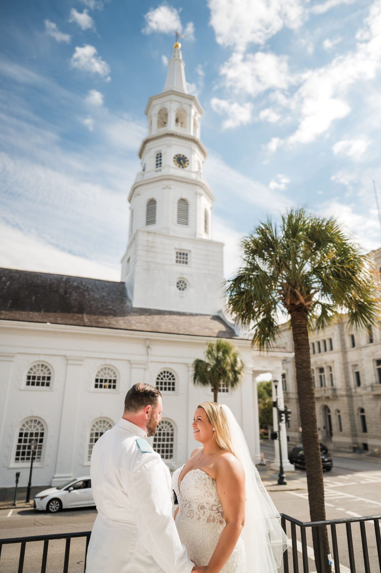 Bride and groom sharing a romantic wedding portrait at The Citadel in Charleston, South Carolina, with the historic white chapel, palm trees, and bright blue skies in the background. Timeless Charleston wedding photography with elegant Southern charm.