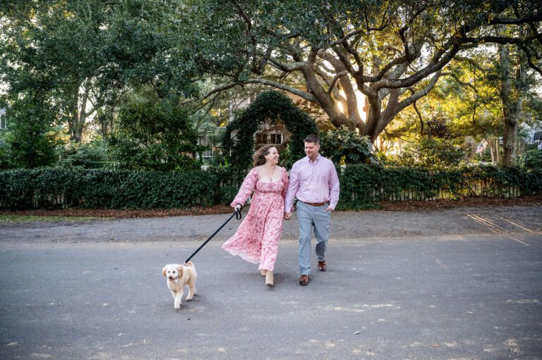 Couple walking hand in hand with their dog during a romantic engagement session beneath moss-covered oak trees in Charleston, South Carolina. Natural Lowcountry engagement photography with warm, timeless style.