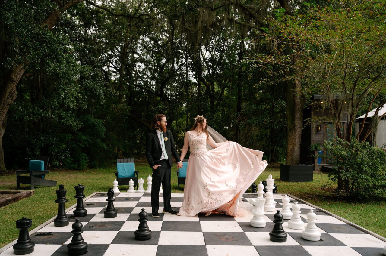 Bride and groom standing on a life-size outdoor chessboard during a romantic wedding portrait at Hart Meadows Ranch. Elegant Charleston area wedding photography featuring a flowing bridal gown and timeless garden scenery.