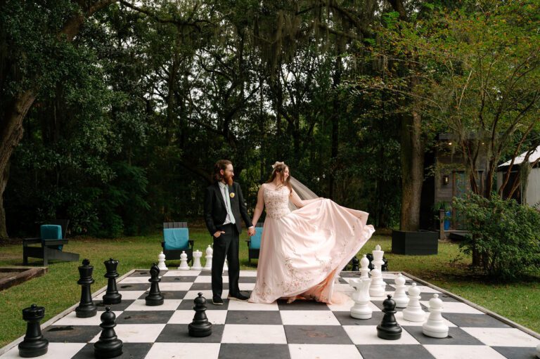 Bride and groom standing on a life-size outdoor chessboard during a romantic wedding portrait at Hart Meadows Ranch. Elegant Charleston area wedding photography featuring a flowing bridal gown and timeless garden scenery.