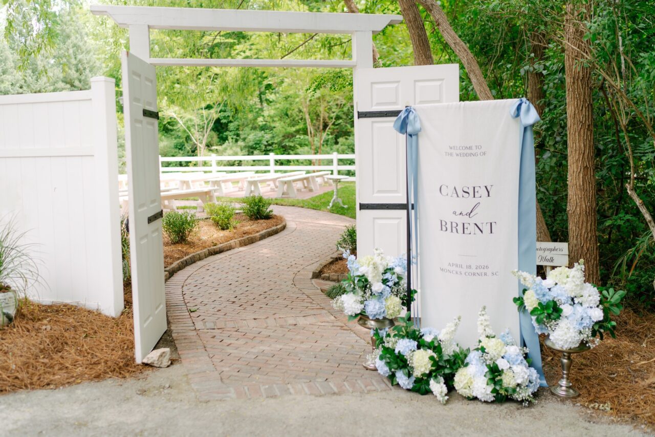 Floral welcome display beside an open white garden entrance and brick ceremony path