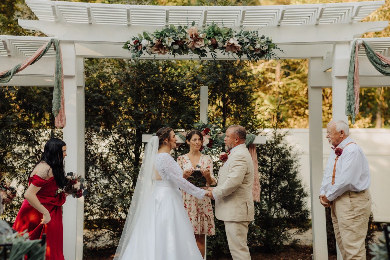 Wedding couple and officiant beneath a white garden pergola decorated with flowers.
