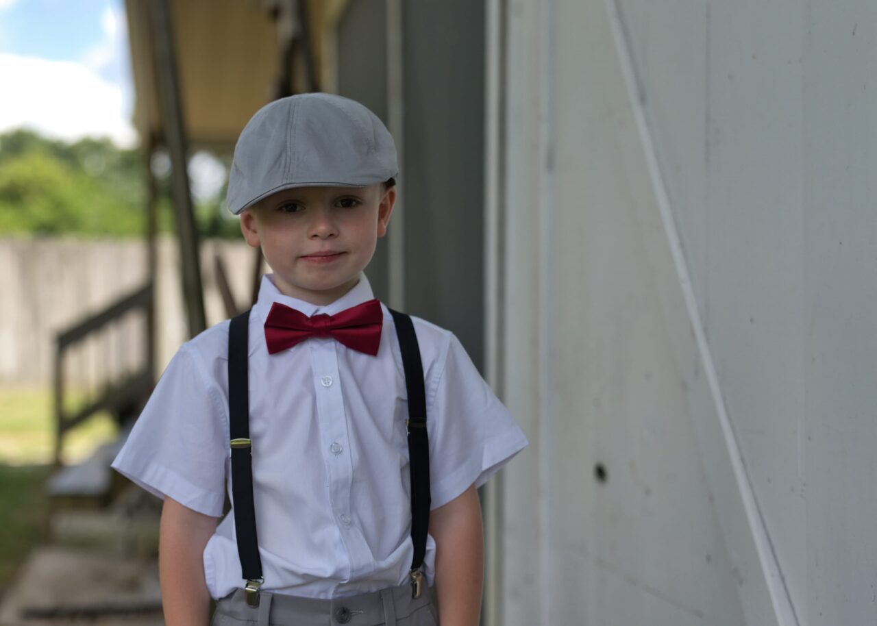 young boy in paper boy hat, bowtie, suspenders, and short sleeve button up.