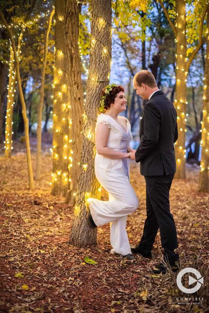 Bride leaning against a Christmas light wrapped tree, looking lovingly at her groom