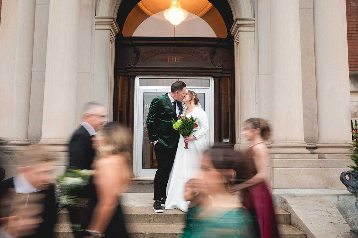 Bride and groom kissing outside of their wedding venue, as their wedding party walks around them. The couple is in focus, but the wedding party has motion blur