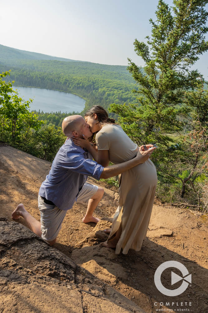 Adam and Nicole kissing while he is on one knee proposes.