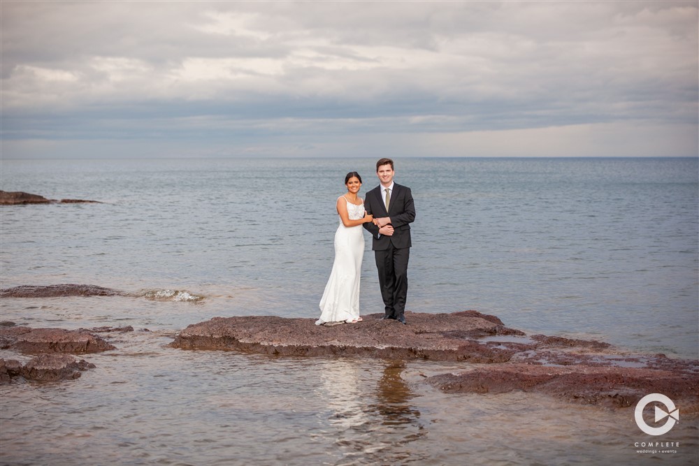 Bride and Groom standing on rock in Lake Superior, photo taken by Complete Weddings + Events Duluth