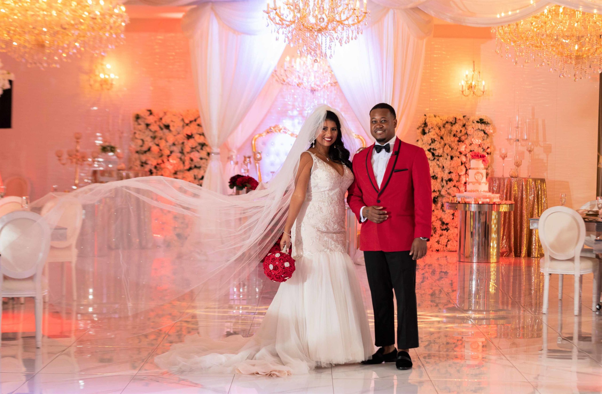 African American bride and groom standing arm and arm. Bride is wearing a gown and groom is wearing a red jacket