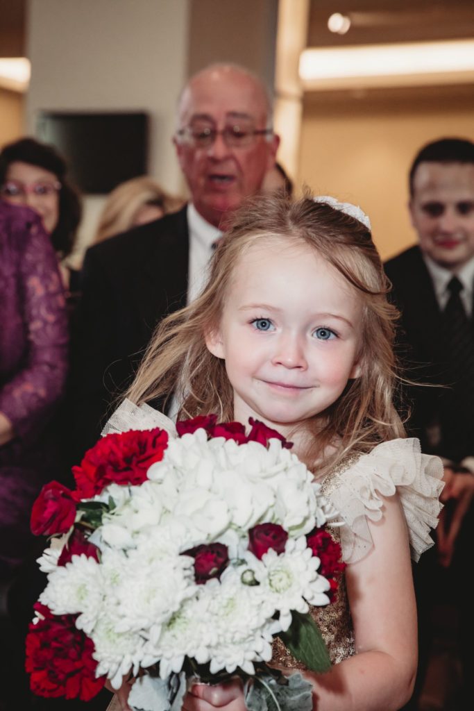 Flower girl smiles at camera holding a giant bouquet and walking down the aisle