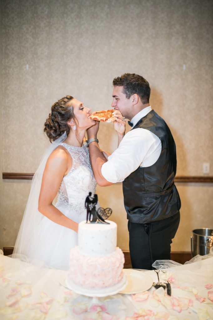 Bride and groom eating pizza while standing behind their wedding cake at Adams Pointe Conference Center in Blue Springs, MO.