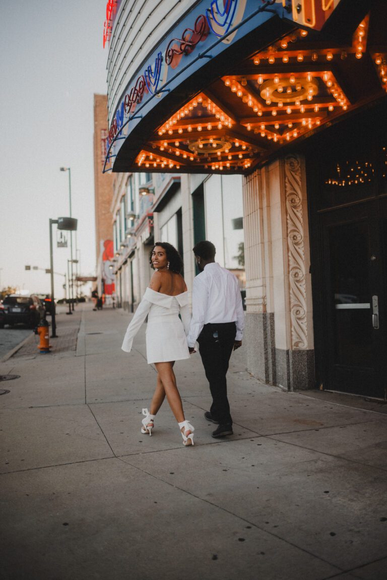 Engaged couple walking down the sidewalk under the Uptown Theater marquee in Kansas City during their engagement session photographed by Kansas City wedding photographer Gabriel with Complete Weddings and Events