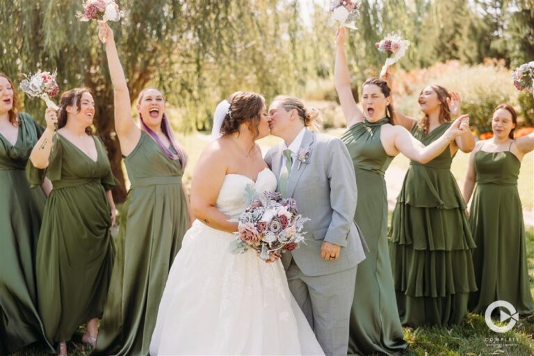 Brides kissing while their wedding party in green dresses cheers behind them in the gardens at The Martin Event Space photographed by Gabriel with Complete Weddings and Events