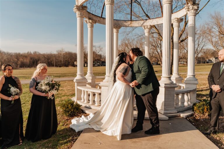 Bride and groom sharing a kiss during their wedding ceremony under the white gazebo at Belvoir Winery photographed by Kansas City wedding photographer Serenity with Complete Weddings and Events