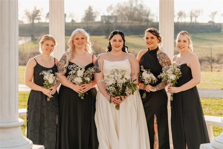 Bride and bridesmaids in black dresses holding white bouquets standing between the gazebo columns at Belvoir Winery wedding venue photographed by Kansas City wedding photographer Serenity