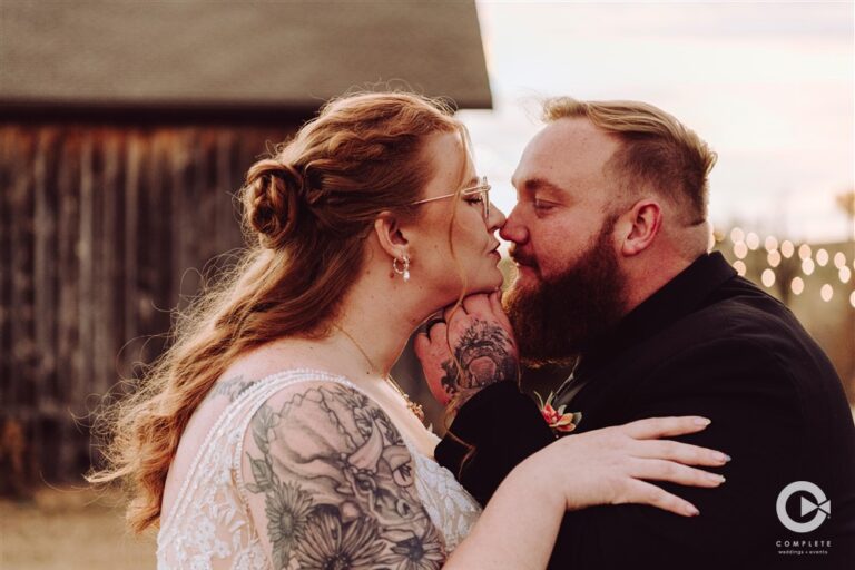Bride and groom sharing a close up kiss at sunset in front of the barn at Backwoods Venue 222 photographed by Serenity with Complete Weddings and Events