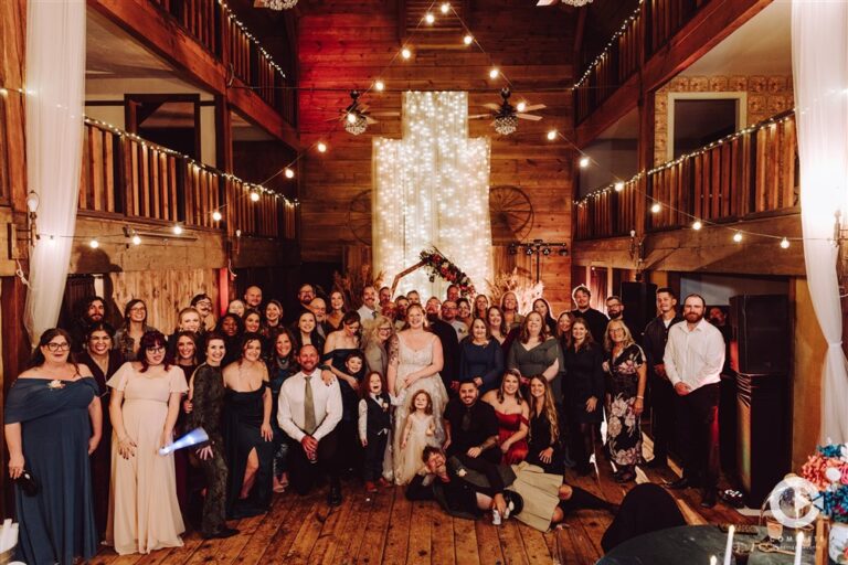 Large wedding guest group photo inside the barn at Backwoods Venue 222 with the bride and groom in the center photographed by Serenity with Complete Weddings and Events
