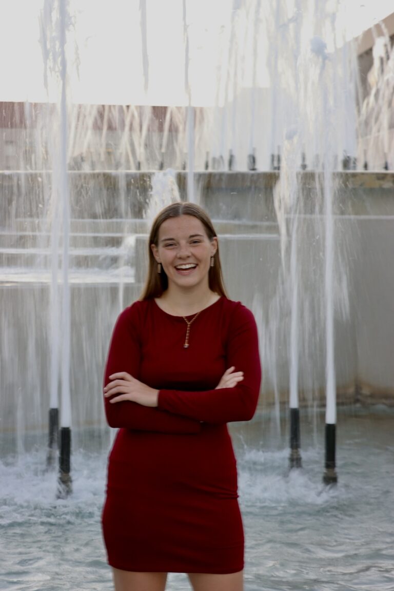 Jill, a Kansas City wedding videographer for Complete Weddings + Events, standing confidently in front of a city fountain in a red dress