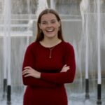 Jill, a Kansas City wedding videographer for Complete Weddings + Events, standing confidently in front of a city fountain in a red dress
