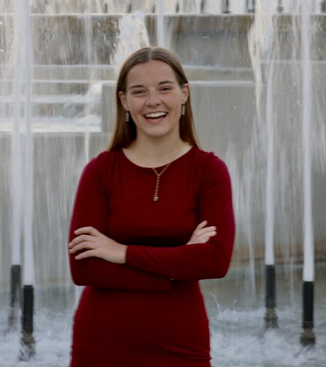 Jill, a Kansas City wedding videographer for Complete Weddings + Events, standing confidently in front of a city fountain in a red dress