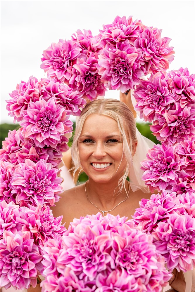 Smiling blonde bride framed by a ring of bright pink dahlia bouquets held around her face for a creative wedding portrait at The Lodge at Ironwoods