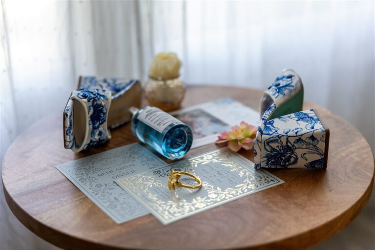 Getting-ready detail flat lay with blue floral napkin holders, perfume, rehearsal dinner invitation, and gold ring on a round wood table at Cedar Valley Forest in De Soto, Kansas