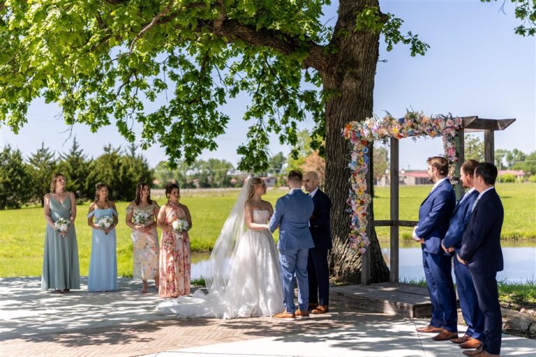 Bride and groom exchanging vows under a large oak tree beside the pond with their wedding party at Cedar Valley Forest in De Soto, Kansas