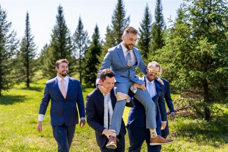 Groom being carried piggyback by groomsmen in navy and blue suits among the evergreen trees at Cedar Valley Forest in De Soto, Kansas