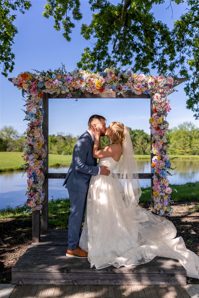 Bride and groom kissing beneath a vibrant wildflower arch beside the pond under a blue sky at Cedar Valley Forest in De Soto, Kansas