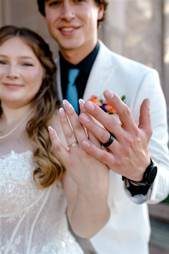 Close-up of a bride's diamond engagement ring and a groom's black wedding band with the smiling couple blurred in the background at a wedding in Ottawa, Kansas