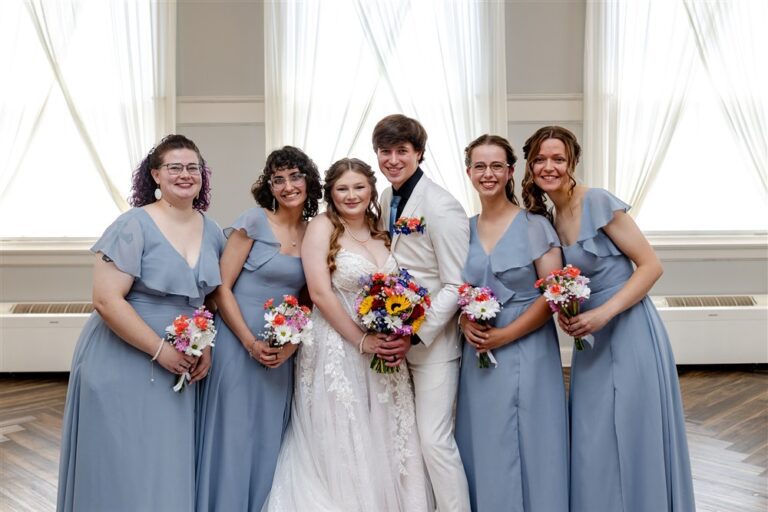 Bride and groom surrounded by four bridesmaids in dusty blue ruffle gowns holding wildflower bouquets at The Lincoln Event Space in Ottawa, Kansas