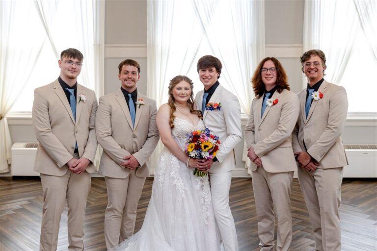 Groom in an ivory tuxedo and four groomsmen in tan suits with dusty blue ties standing with the bride in the ballroom at The Lincoln Event Space in Ottawa, Kansas