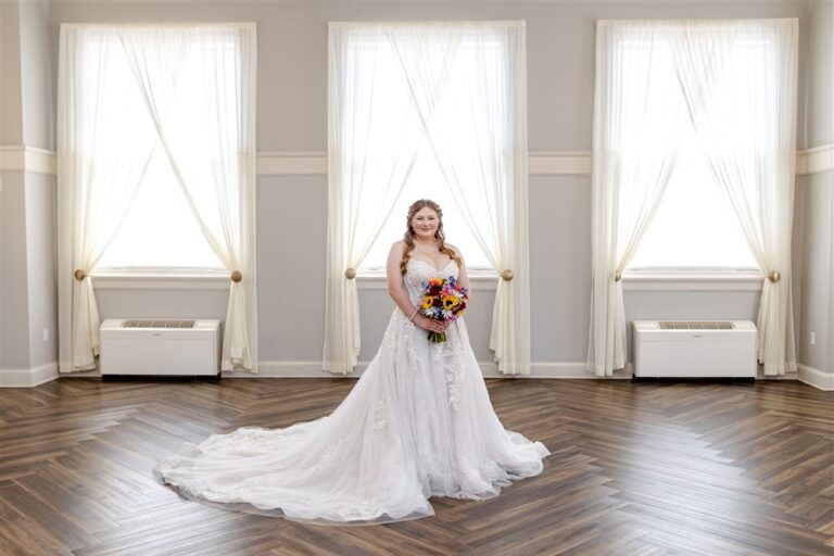 Bride in a lace A-line wedding gown with a long train holding a sunflower and wildflower bouquet in the sunlit ballroom at The Lincoln Event Space in Ottawa, Kansas