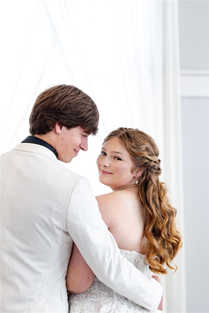 Bride in a strapless lace gown looking back over her shoulder while her groom holds her, backlit by a sheer white curtain at a wedding in Ottawa, Kansas
