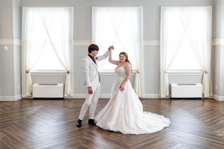 Groom twirling his bride in her lace ball gown on the herringbone wood dance floor beneath tall draped windows at The Lincoln Event Space in Ottawa, Kansas