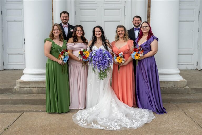 Bride with her bridesmaids and groomsmen posing between white columns on the front steps of First Baptist Church in Richmond, Missouri