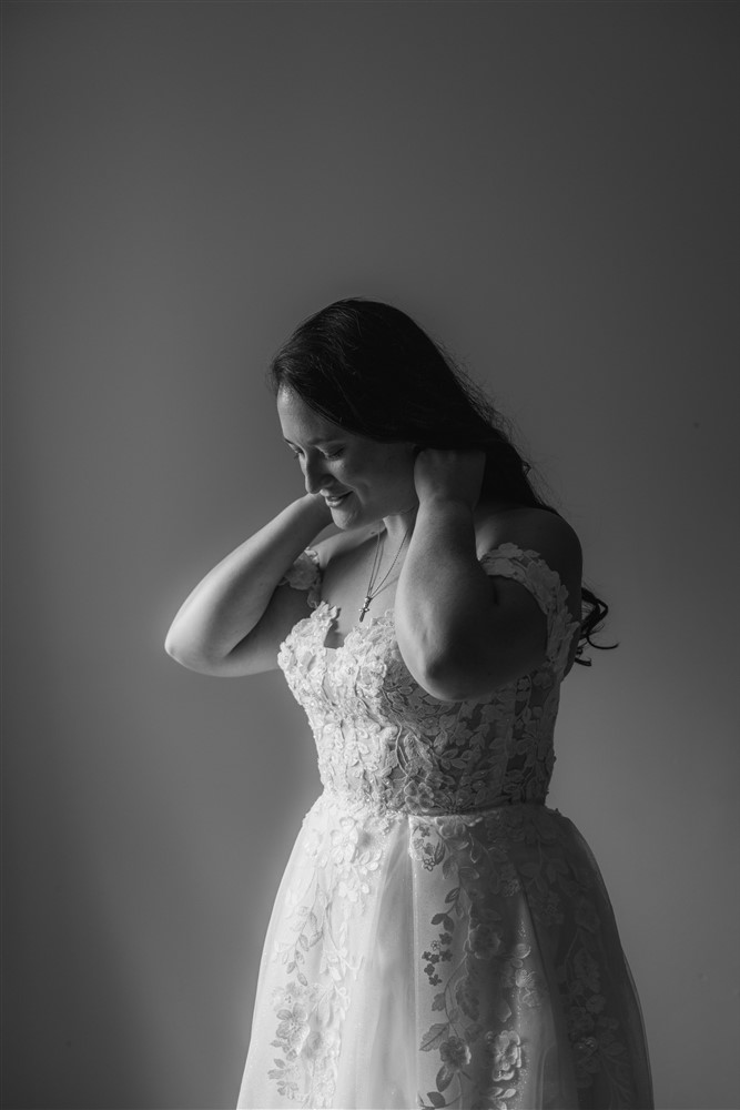 Black and white portrait of a bride in a floral appliqué gown looking down and lifting her hair against a plain wall at a Richmond, Missouri wedding
