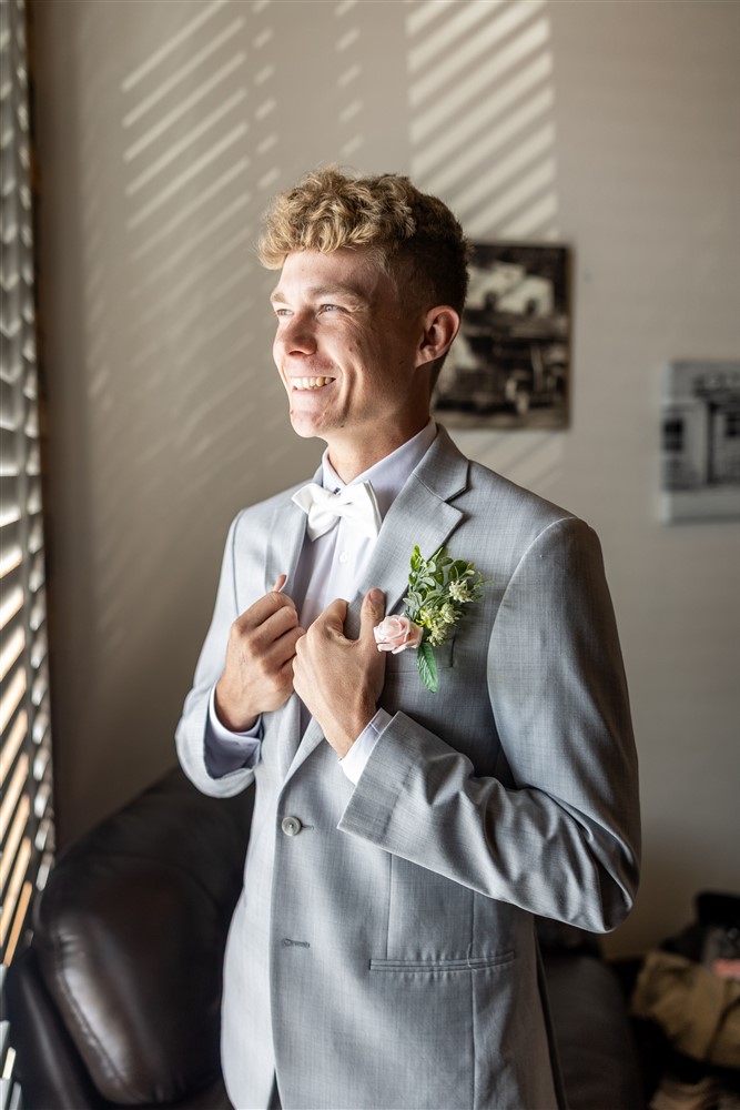 Groom in a light grey suit adjusts his jacket in window light while getting ready for his wedding