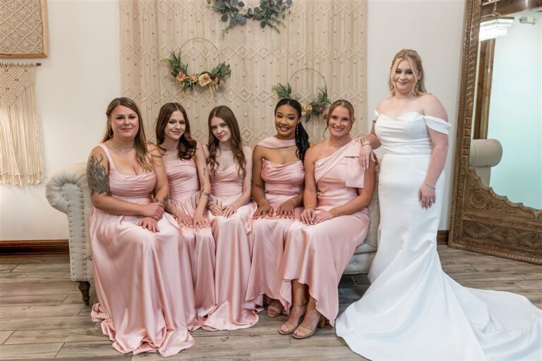 Five bridesmaids sit on a couch in the bridal suite while the bride stands beside a macrame backdrop
