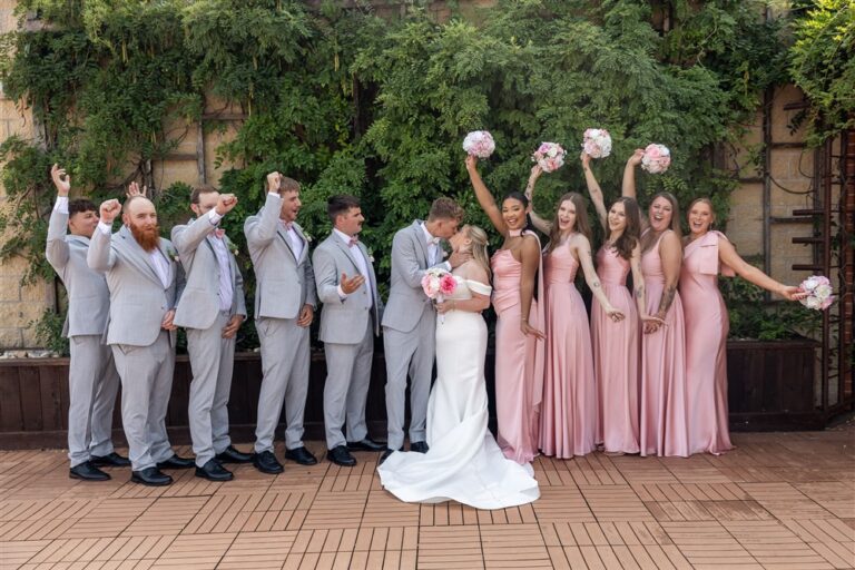 Full wedding party cheers with arms raised as the bride and groom kiss at The Stanley in Lee's Summit