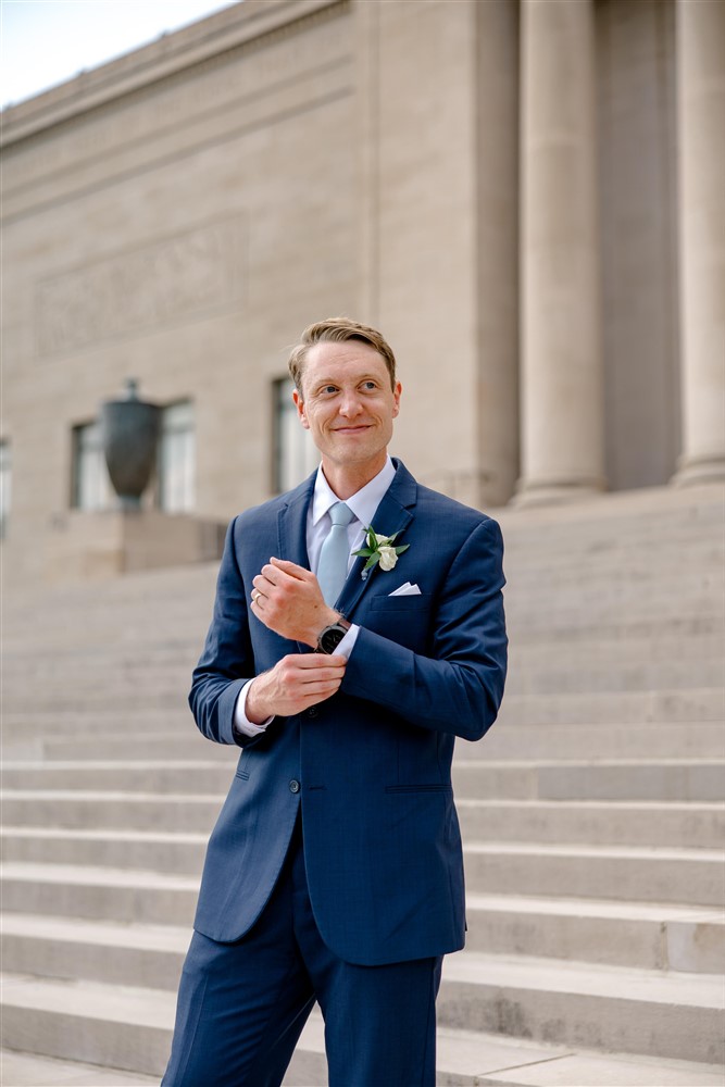 Groom in a navy suit and light blue tie adjusting his cuff on the limestone steps of the Nelson-Atkins Museum of Art in Kansas City