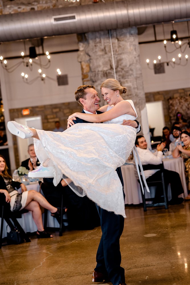 Groom lifting his bride in his arms as they laugh during their first dance at a Stone Manor on 79th wedding reception in Overland Park, Kansas