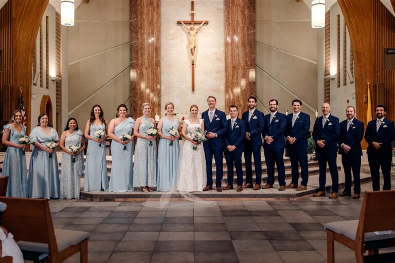 Bride, groom and their full wedding party standing at the altar of a Catholic church beneath a large crucifix after the ceremony