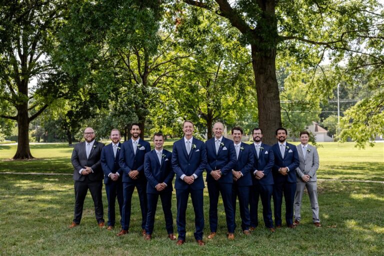 Ten groomsmen in navy and grey suits with white rose boutonnieres lined up for a formal portrait under shade trees