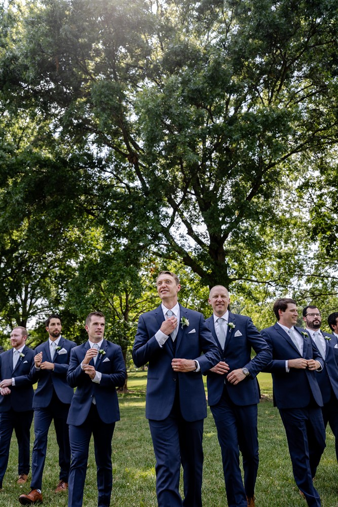 Groom and his groomsmen in navy suits walking together and adjusting their ties under summer trees at a Kansas City wedding
