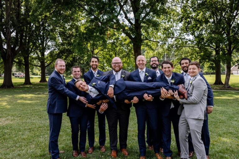 Groomsmen in navy suits holding the groom horizontally overhead and laughing during outdoor wedding party photos