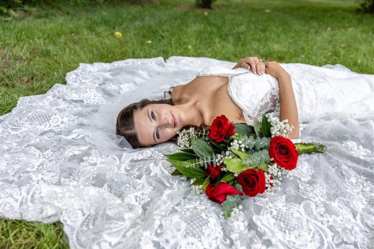 Bride lies in the grass with her lace gown fanned around her and a red rose bouquet resting at her shoulder in Blue Springs, Missouri