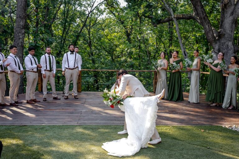 Groom dips his bride for a kiss on the lawn while the wedding party applauds from the wooden deck at a Blue Springs, Missouri wedding