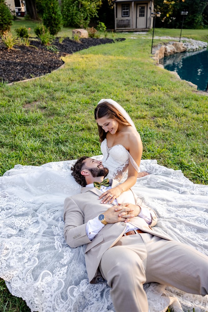 Groom lies across his bride's lap on the lawn beside a pond during golden hour wedding portraits in Blue Springs, Missouri