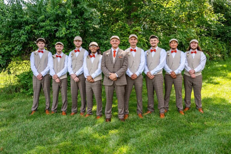 Groom and eight groomsmen in tan vests, brown plaid trousers, burnt orange bow ties and flat caps lined up at Tobacco Barn Farm
